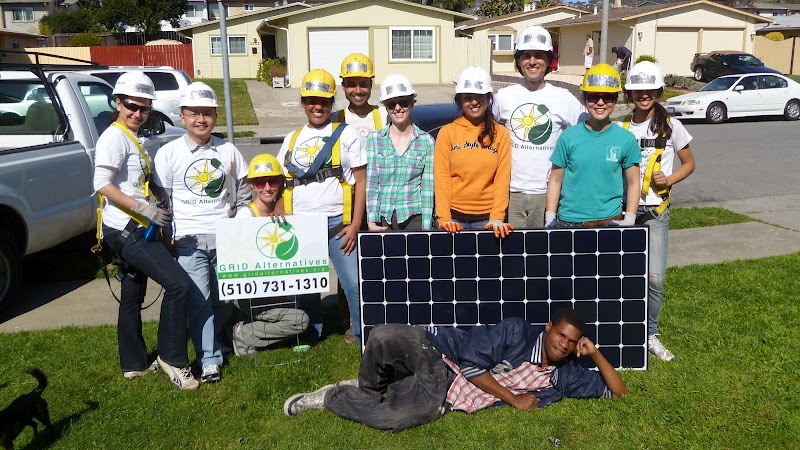Group of volunteers in hard hats holding solar panel, standing on lawn with banner displaying phone number (510) 731-1310