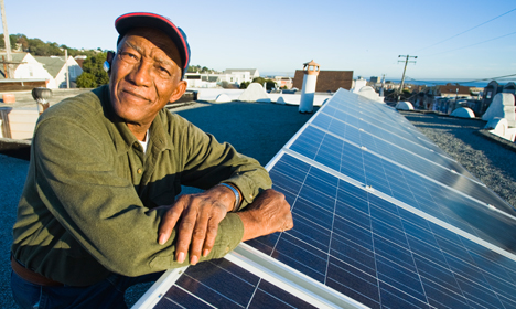 Man in green jacket smiling while leaning on solar panel on rooftop