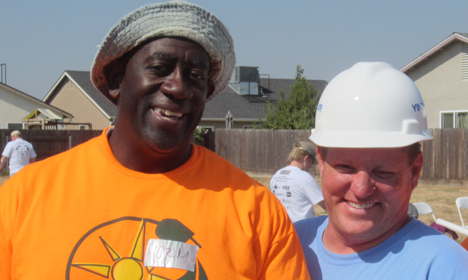 Two men smiling at outdoor event; one wearing orange shirt and gray hat, other in blue shirt and white hard hat