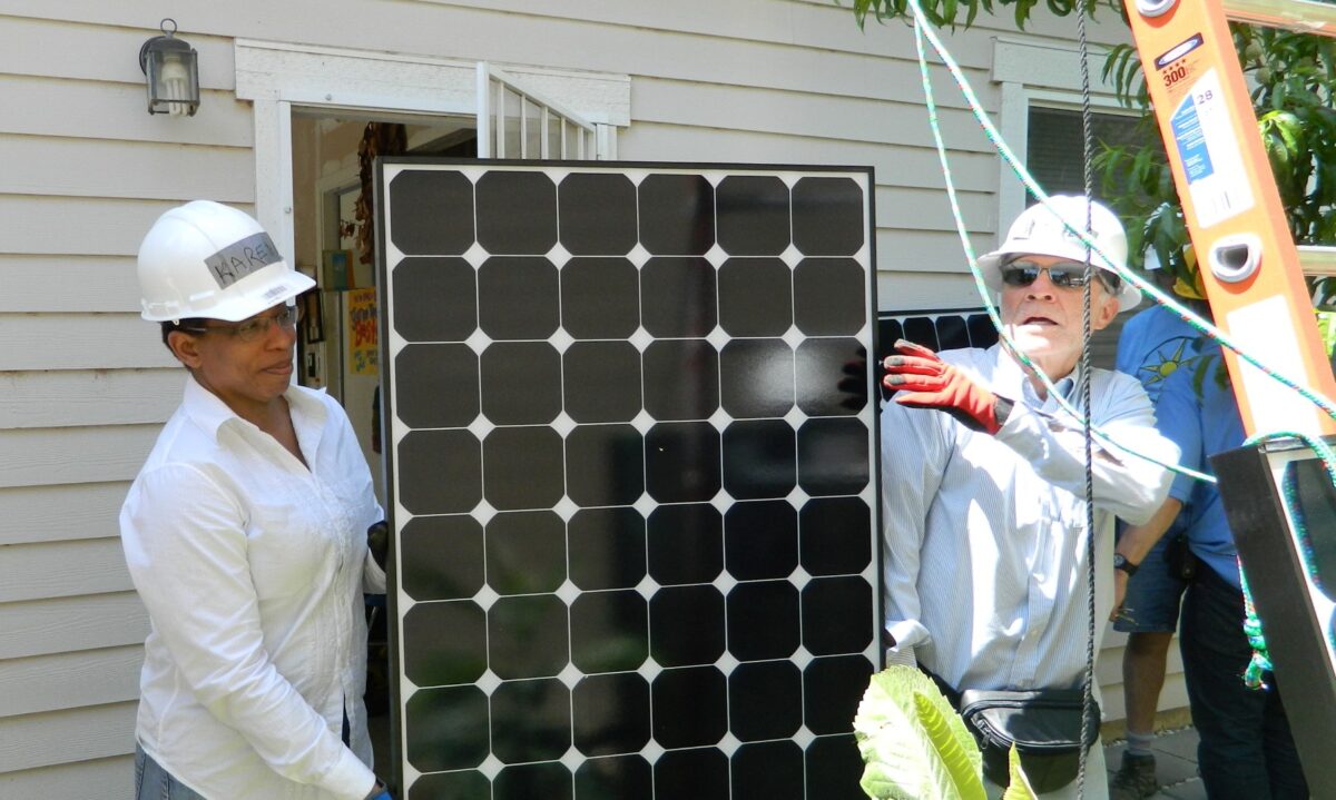 Two workers in white hard hats installing a solar panel on a residential house exterior