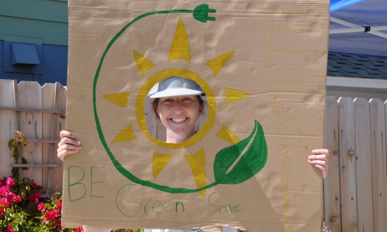 Person smiling through sun-shaped cutout on cardboard sign reading "Be Green Save