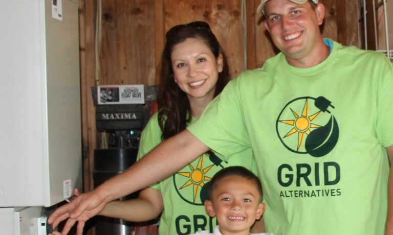 Three people wearing green Grid Alternatives shirts smile together in front of solar equipment indoors