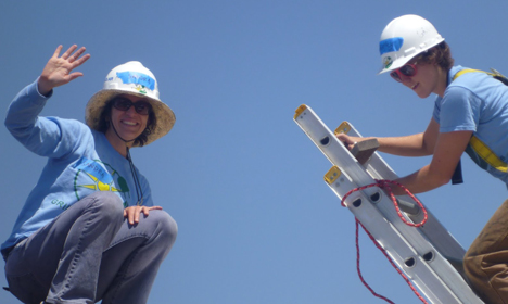Two workers in hard hats and blue shirts celebrate outdoors with a ladder and tools visible