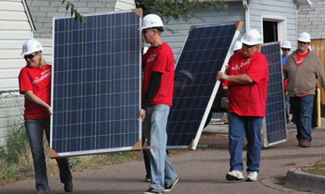 Group of volunteers in red shirts and hard hats carrying large solar panels during an installation project
