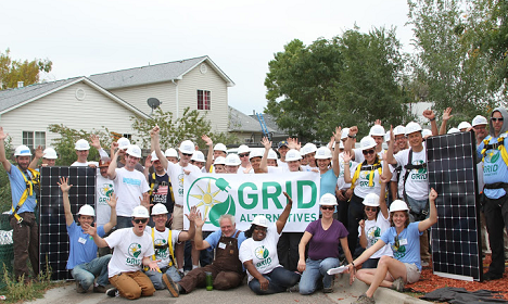 Large group wearing GRID initiative shirts and white hats pose with solar panels at community event