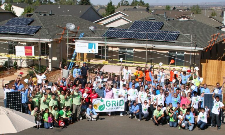 Large group holding GRID Alternatives banner in front of solar panel-equipped house during community event
