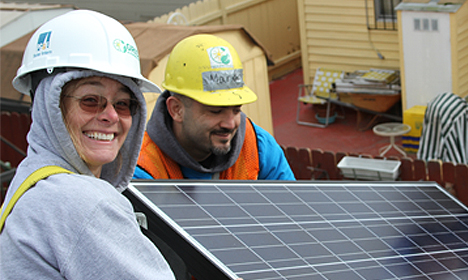 Two workers in hard hats and safety vests smiling while holding a solar panel at a job site