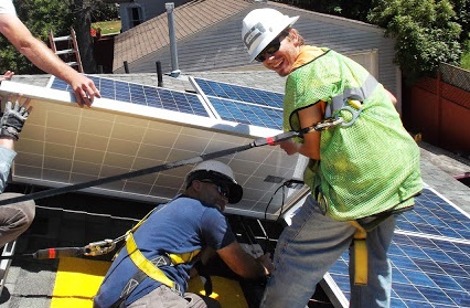 Two workers installing solar panels on a residential roof, one in yellow safety vest guiding placement