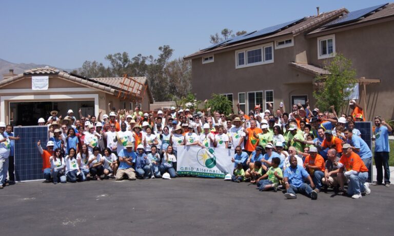 Large group of volunteers in safety vests and hard hats gathered in driveway holding GRID Alternatives banner in front of solar-installed house