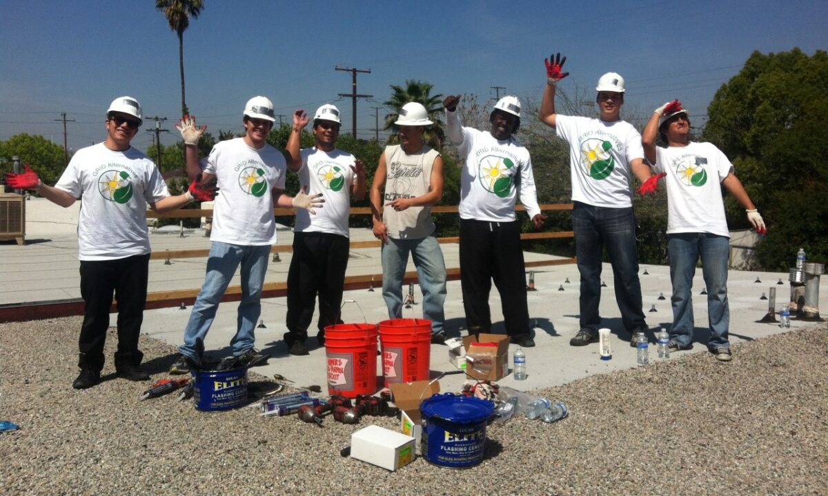 Seven construction workers in white hard hats and matching t-shirts pose on a rooftop with solar installation materials and tools