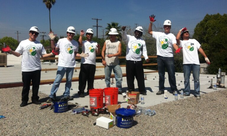 Seven construction workers in white hard hats and matching t-shirts pose on a rooftop with solar installation materials and tools