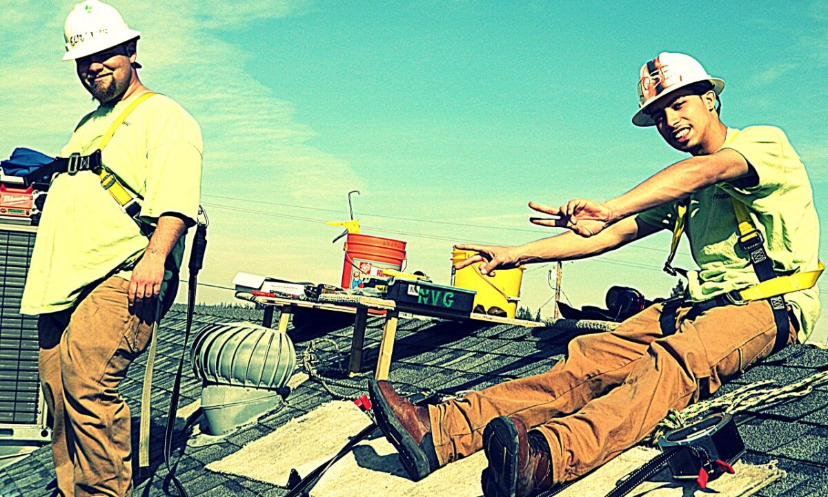 Two construction workers in yellow shirts and hard hats installing roofing materials on a sunny day