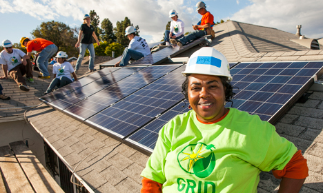 Worker in green GRID shirt smiles on residential rooftop with solar panels and installation team visible