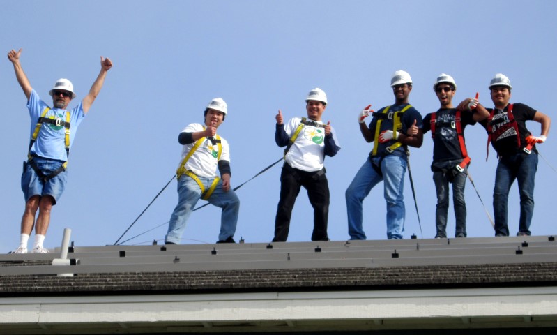 Seven construction workers in hard hats stand on a roof against a clear blue sky, posing enthusiastically with ropes and tools