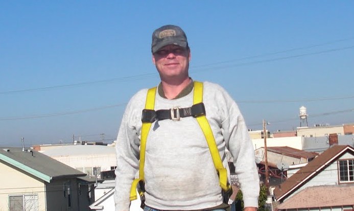Man wearing safety harness and cap on rooftop with industrial buildings in background