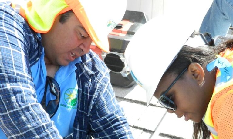 Two young interns in safety gear and hard hats examine solar panel equipment during a work training session