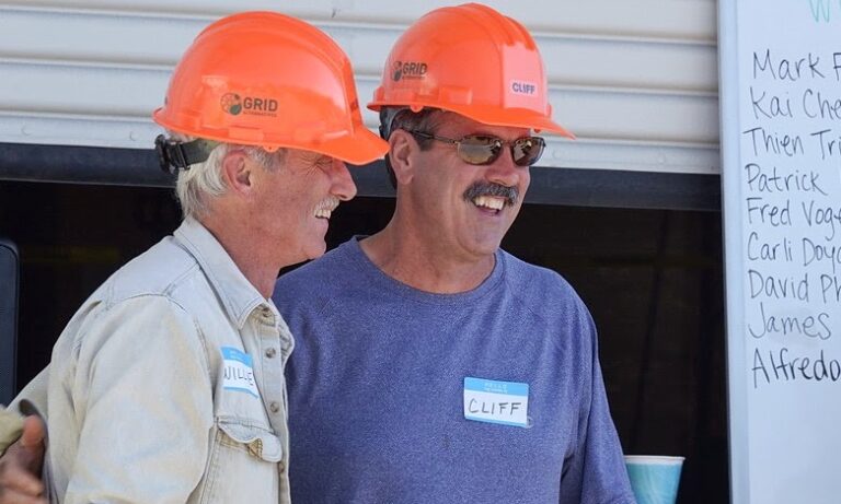 Two men wearing orange GRID hard hats smile together at a volunteer event, with a list of volunteer names visible on a whiteboard behind them