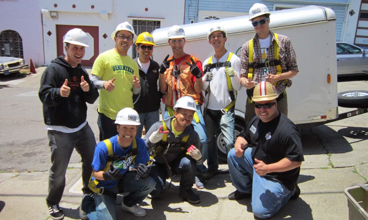 Construction team of nine workers in hard hats and safety vests giving thumbs up by a trailer during Genentech volunteer project