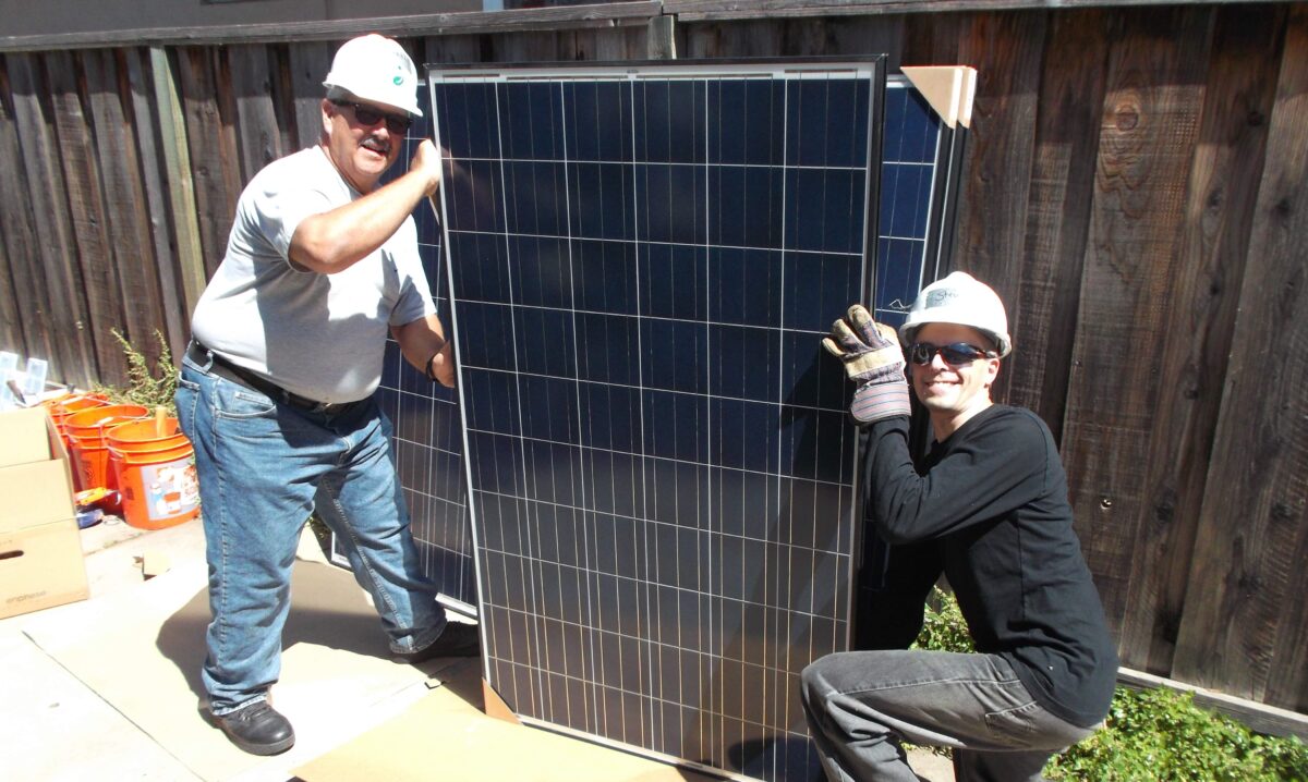 Two solar installation workers holding a large dark blue solar panel outdoors on a sunny day