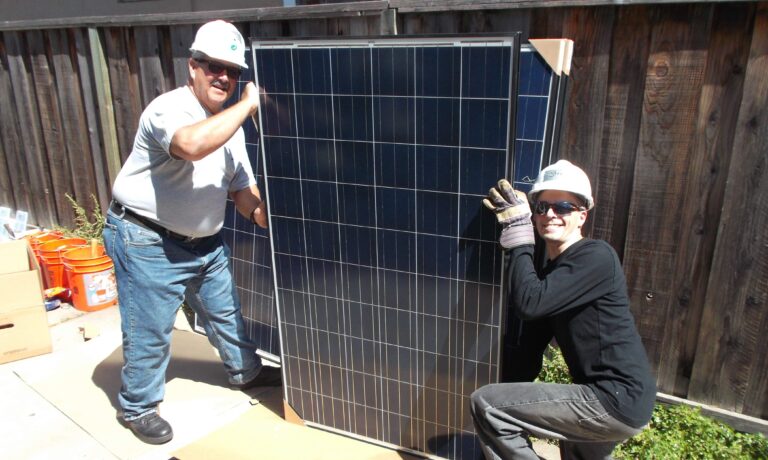 Two solar installation workers holding a large dark blue solar panel outdoors on a sunny day