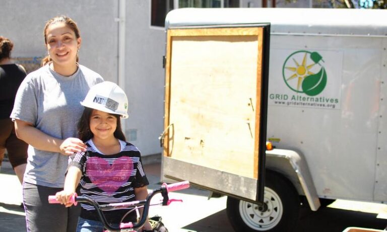 Two young girls smile at a GRID Alternatives solar energy trailer during a community event