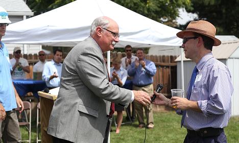 Two men shake hands at an outdoor event under a white tent with spectators in background