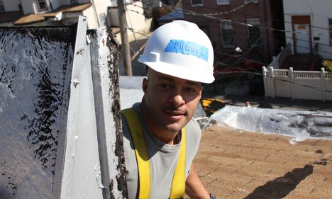 Construction worker in white hard hat and yellow safety vest on rooftop with buildings in background