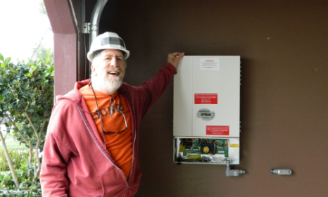 Bearded man in hard hat and red jacket pointing to a wall-mounted electrical panel outdoors