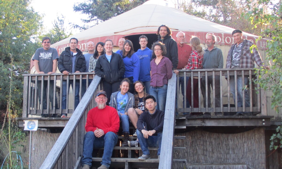 Group of approximately 18 people posed on wooden deck steps under a tent during an outdoor event or team gathering