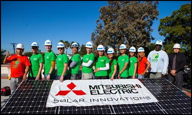 Group of people in hard hats and green shirts standing behind Mitsubishi Solar Innovations banner with solar panel