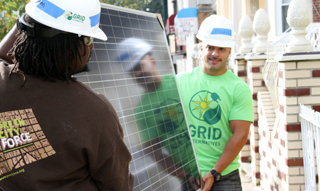 Two workers in hard hats examining a solar panel on a rooftop, one wearing a green GRID shirt
