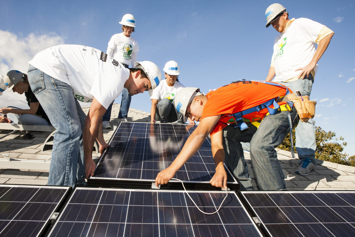 Team of volunteers in hard hats installing solar panels on a rooftop under clear blue sky