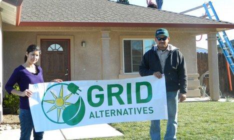 Two people holding Grid Alternatives banner in front of residential home under construction