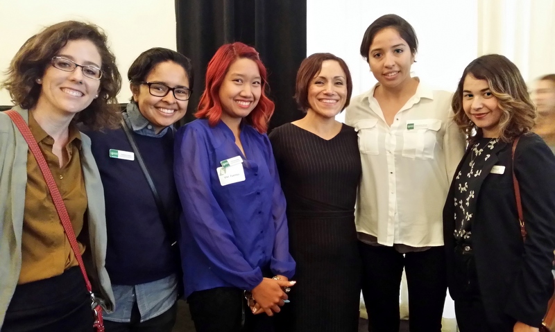 Six women smiling at camera in professional setting, wearing name badges and casual business attire