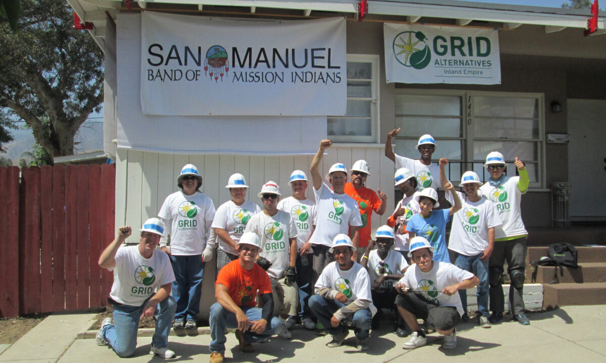 Group of volunteers in hard hats and GRID Alternatives shirts pose in front of San Manuel Band of Mission Indians building