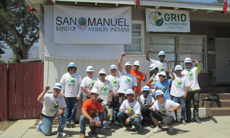 Group of volunteers in hard hats and GRID Alternatives shirts pose in front of San Manuel Band of Mission Indians building