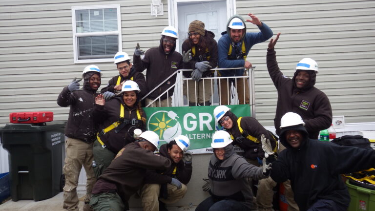 Group of workers in hard hats and safety vests posing together outside a house with Green City Force banner