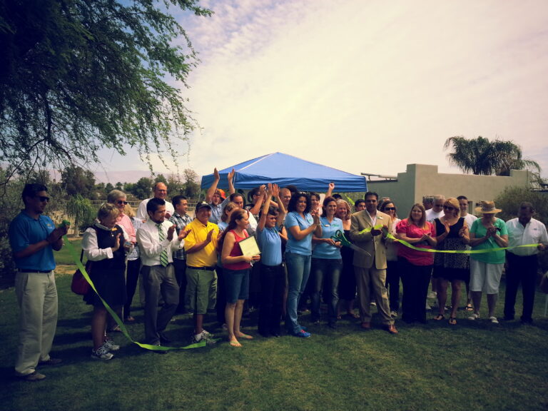Group of people holding a large green ribbon for a ribbon-cutting ceremony outdoors under a blue tent