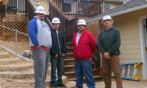 Four construction workers in hard hats stand on residential deck stairs during a worksite visit