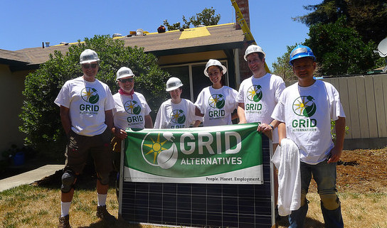 Six volunteers in white hard hats and GRID Alternatives t-shirts hold a green GRID Alternatives banner with solar panel installation in background