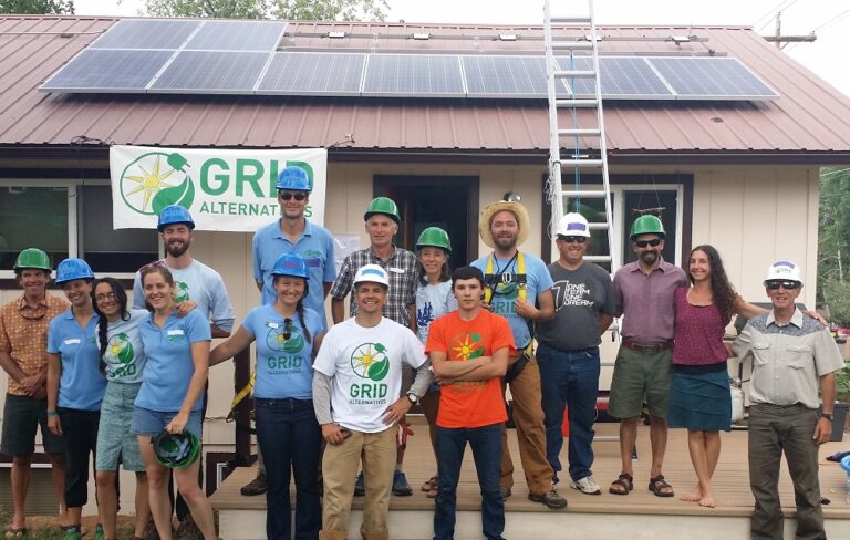 Group photo of GRID Alternatives team members and volunteers wearing hard hats in front of a building with solar panels on the roof