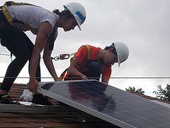 Two workers installing solar panels on a residential roof wearing hard hats and safety gear