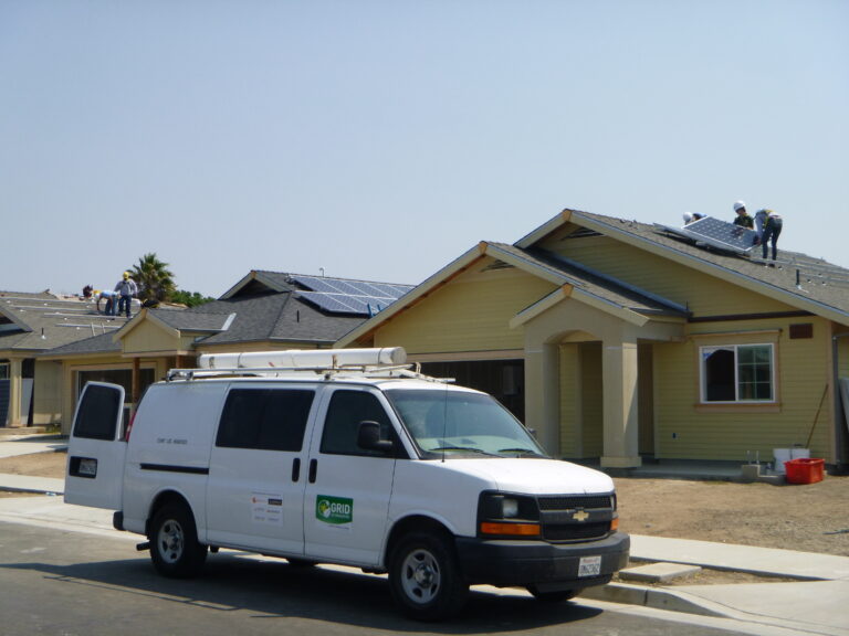 White van labeled "Grid" parked in front of yellow house with solar panels being installed on roof by workers
