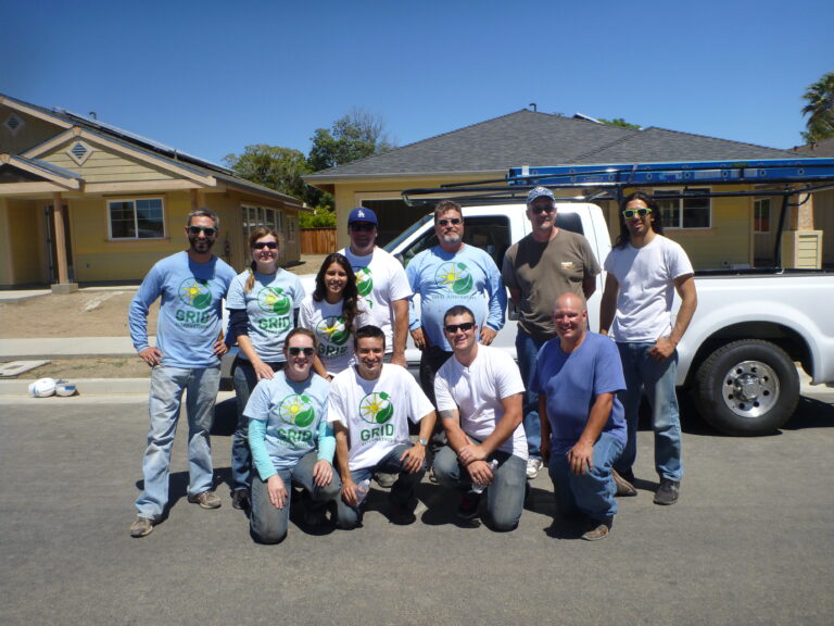 Volunteer team in GRID t-shirts poses in front of yellow house with white pickup truck on sunny day