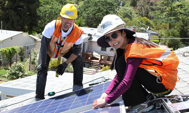 Two workers installing solar panels on a roof, wearing safety helmets and orange vests