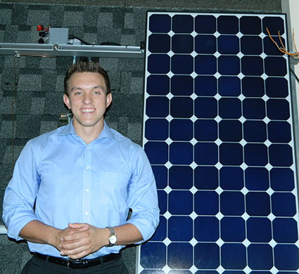 Man in light blue shirt standing next to large solar panel on green shingle roof