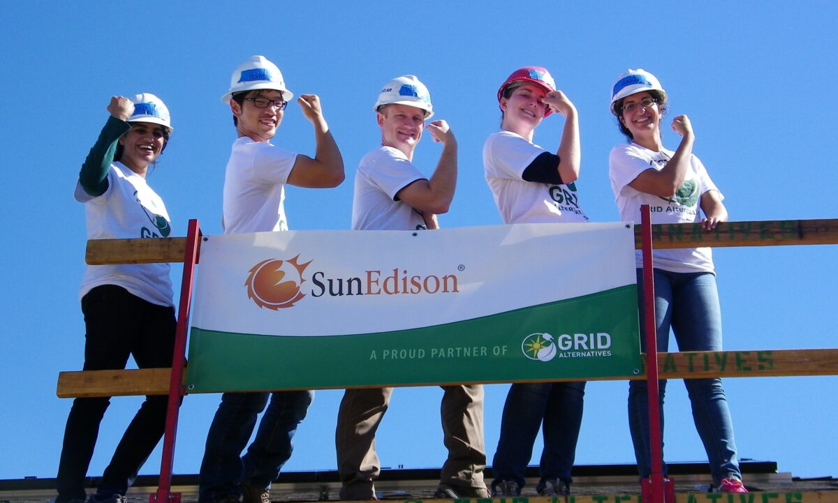 Five construction workers in hard hats posing with a SunEdison banner and GRID Alternatives logo against blue sky