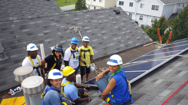 Students in hard hats and safety gear work together installing solar panels on a residential roof