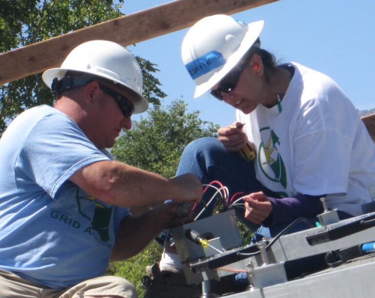 Two workers in hard hats installing electrical wiring on utility equipment outdoors