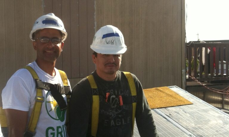 Two construction workers in hard hats and safety vests stand on a rooftop at City College of San Francisco